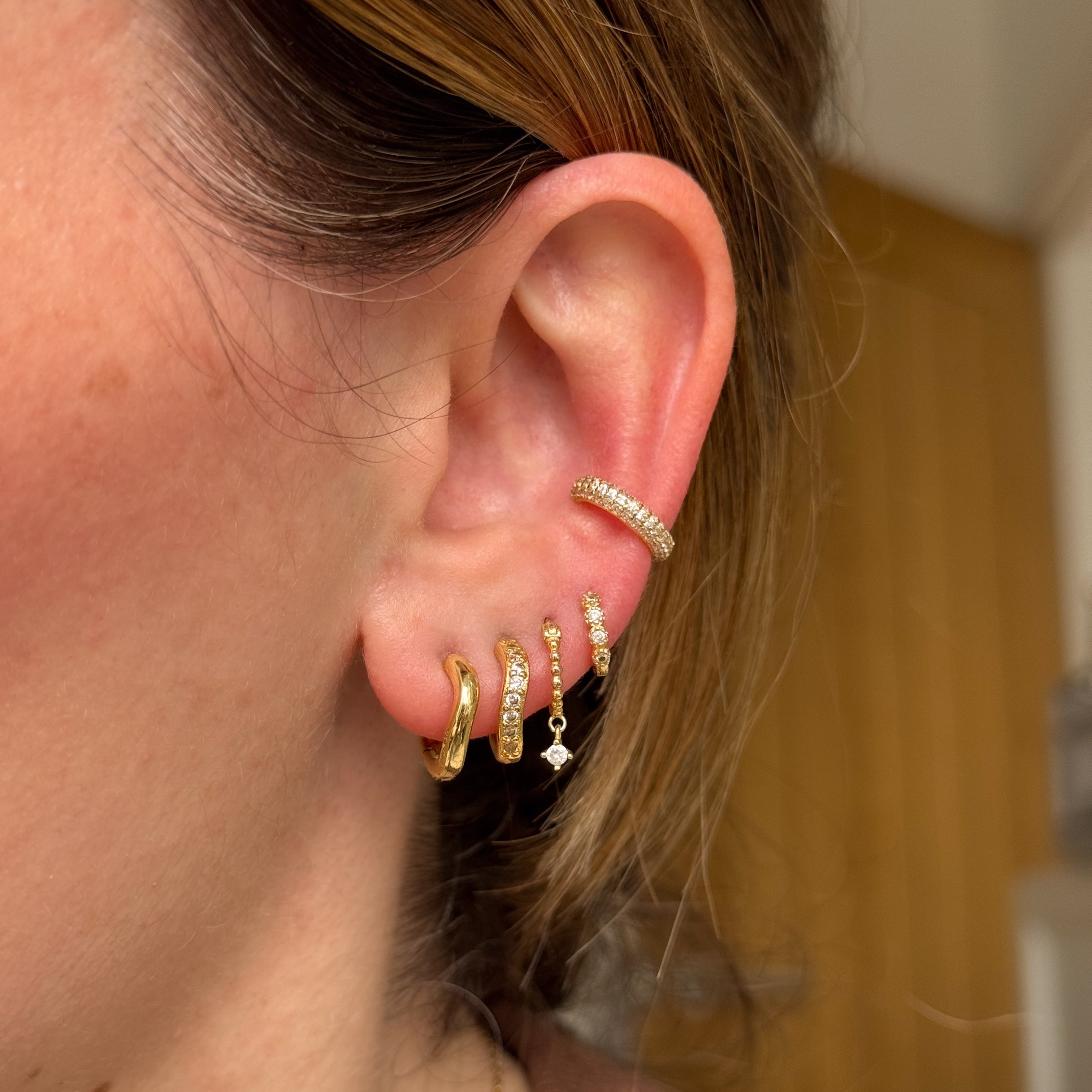 Close-up of an ear wearing gold dainty huggie hoop earrings with small gemstones and an ear cuff.