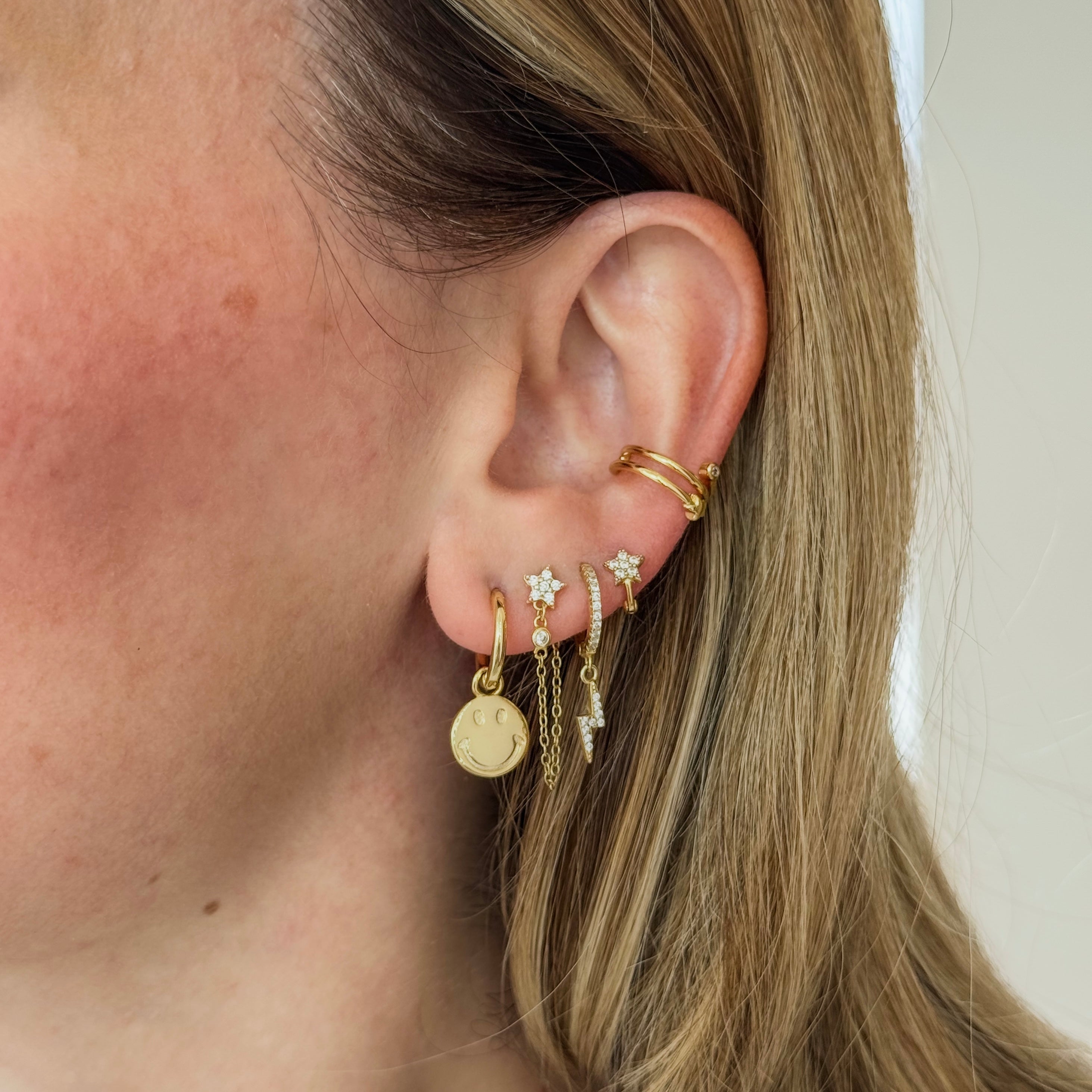 Close-up of an ear with multiple gold huggie style earrings and ear cuff on a neutral background