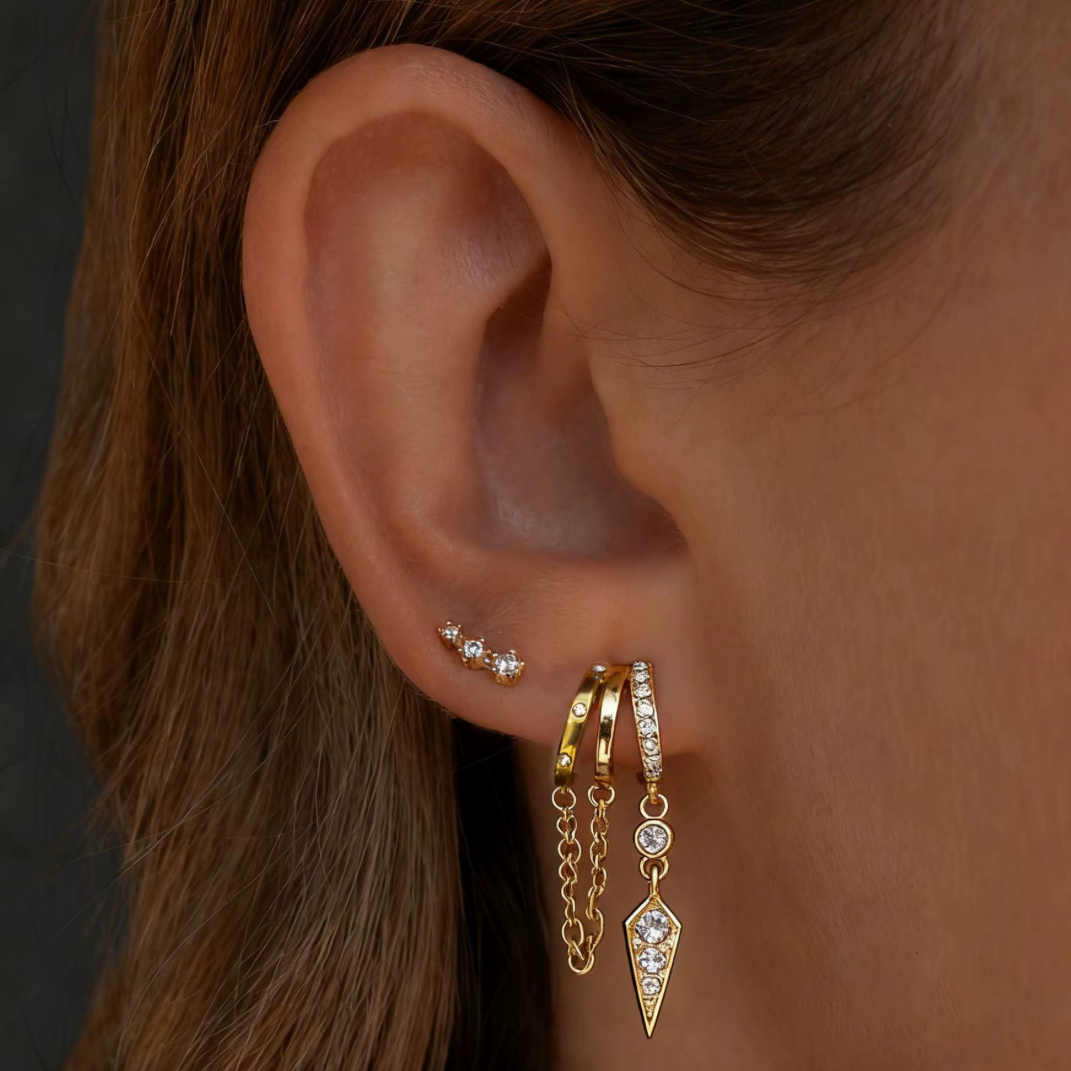 Close-up of an ear wearing gold hoop earrings with chain and diamond accents.