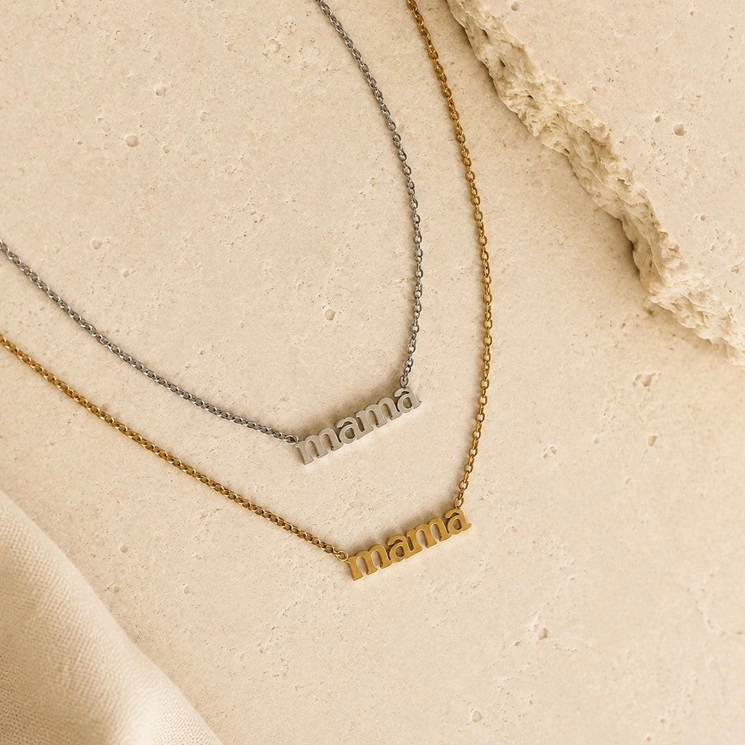 Two name necklaces spelling 'mama' in lowercase letters, one in gold and one in silver, displayed on a textured beige surface with a piece of natural stone and soft fabric.