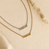 Two name necklaces spelling 'mama' in lowercase letters, one in gold and one in silver, displayed on a textured beige surface with a piece of natural stone and soft fabric.