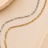 Two paperclip-link chains, one gold and one silver, arranged in parallel curves on a warm beige background with textured stone edges.