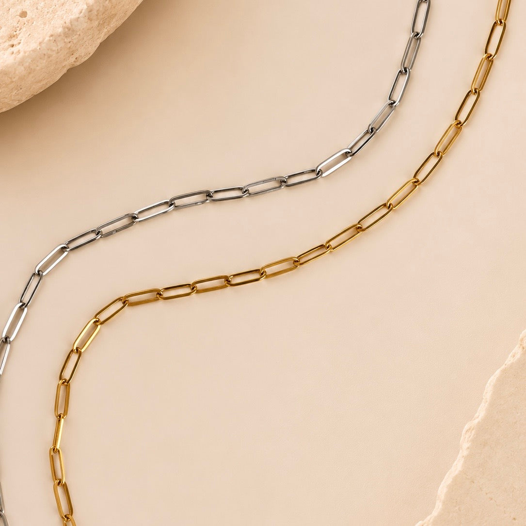 Two paperclip-link chains, one gold and one silver, arranged in parallel curves on a warm beige background with textured stone edges.
