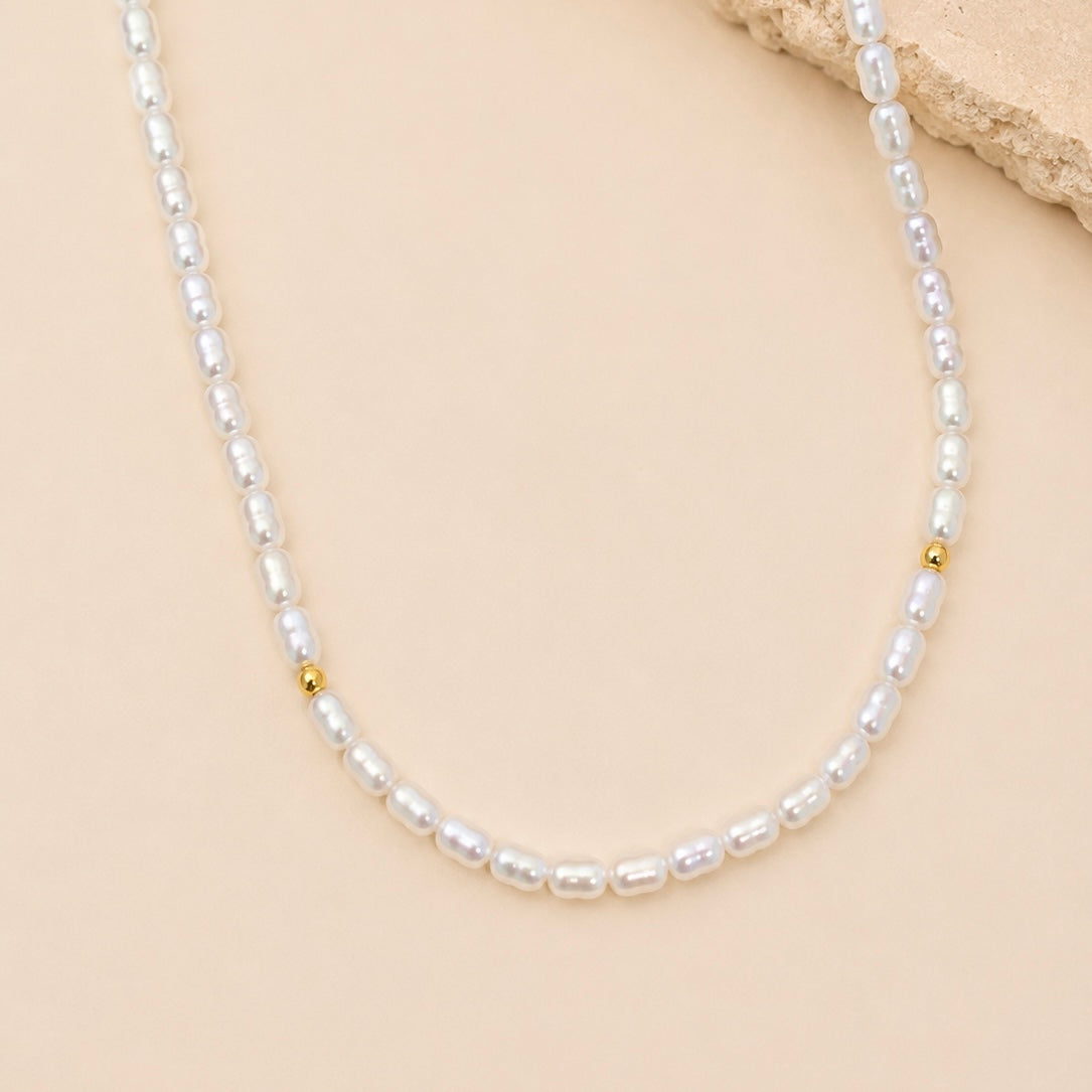 A delicate necklace made of small white baroque pearls with small gold spacer beads, displayed on a beige background with a textured stone prop.