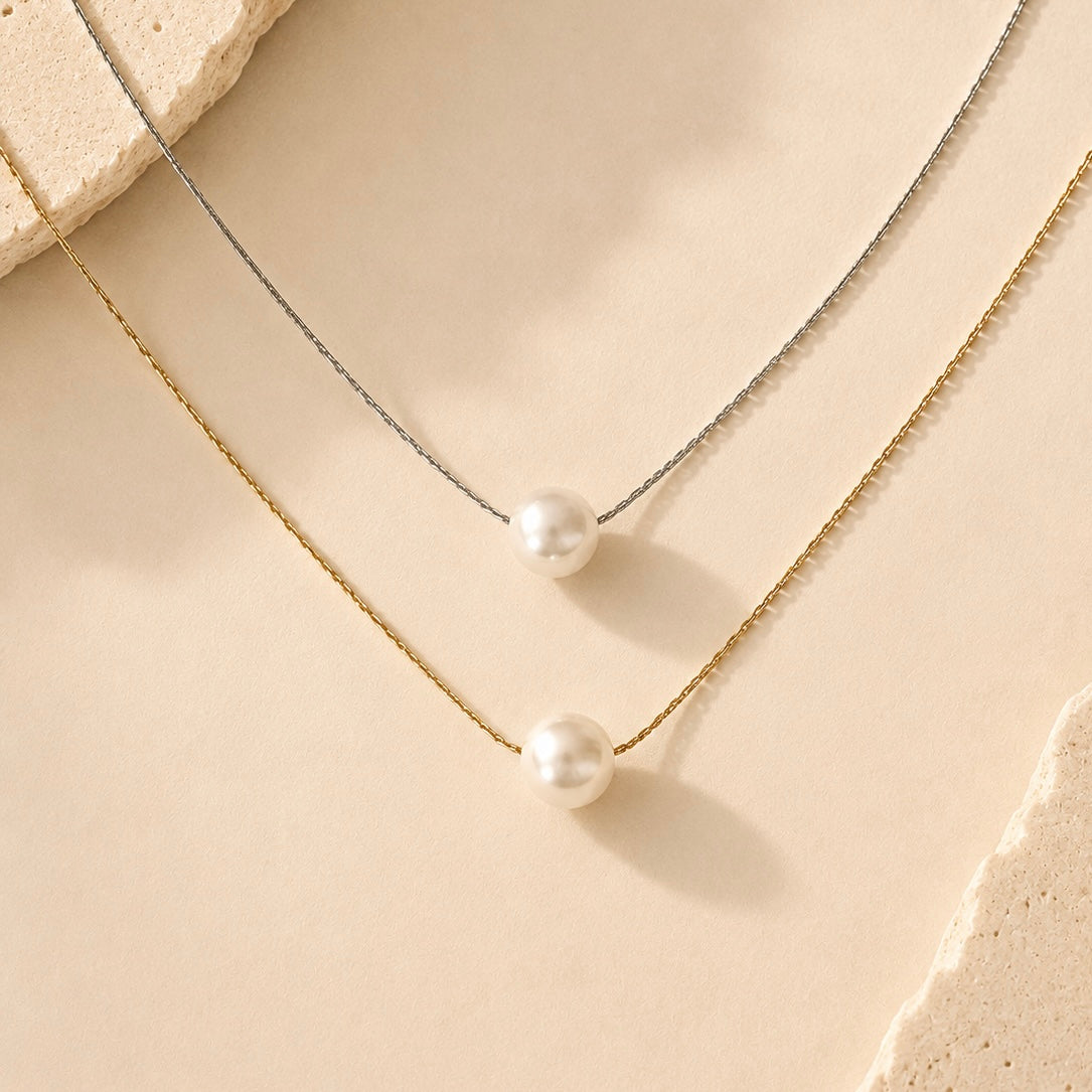 Two delicate necklace chains with white pearl pendants, one in gold-tone metal and one in silver-tone metal, arranged on a warm beige textured surface.