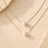 Two delicate necklace chains with white pearl pendants, one in gold-tone metal and one in silver-tone metal, arranged on a warm beige textured surface.
