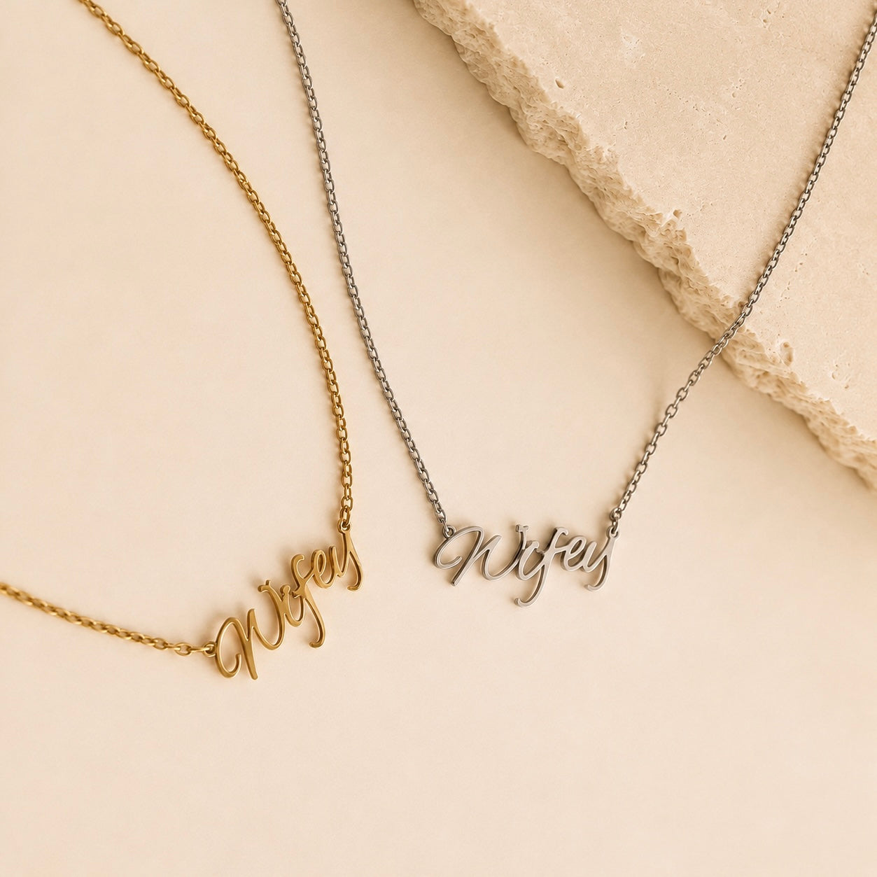 Two name necklaces with cursive script pendants, one in gold and one in silver, displayed on a light beige background with a textured stone prop.