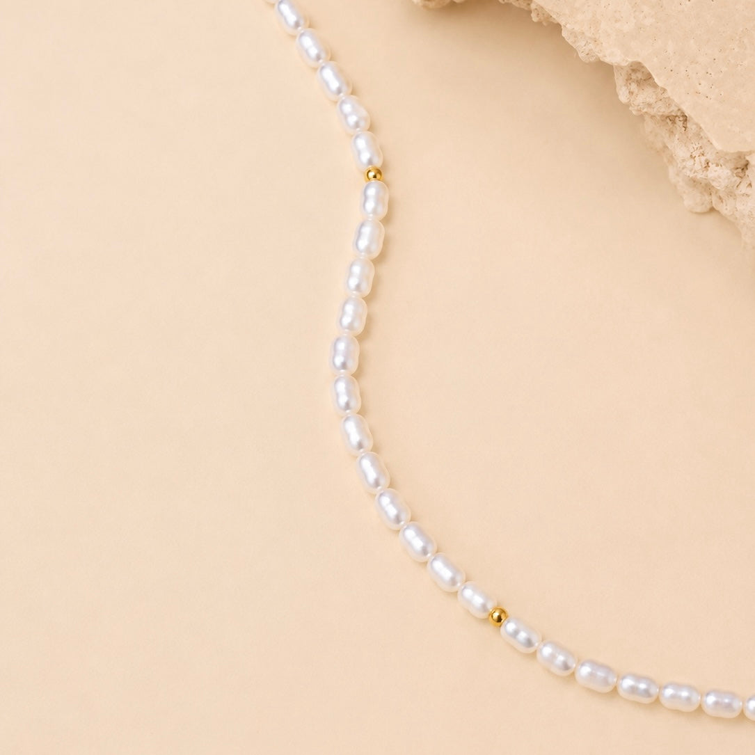 A delicate strand of white baroque pearls separated by small gold spacer beads, laid out on a warm beige background with a textured stone prop in the top right corner.
