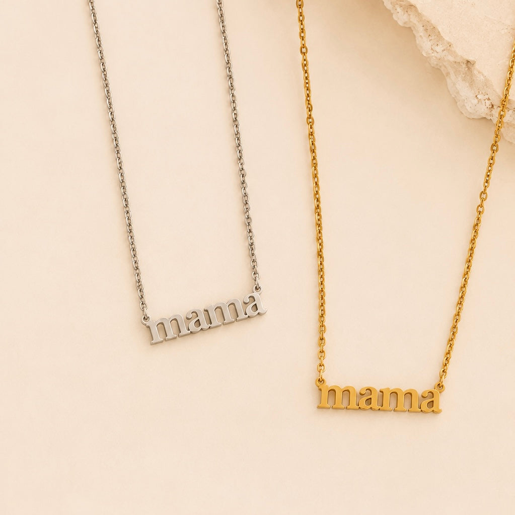 Two name necklaces with the word 'mama' in lowercase letters, one in silver and one in gold, displayed on a light cream background with a decorative white stone prop.