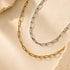 Two paperclip chain necklaces in gold and silver tones, arranged on a warm beige textured surface with stone accents.