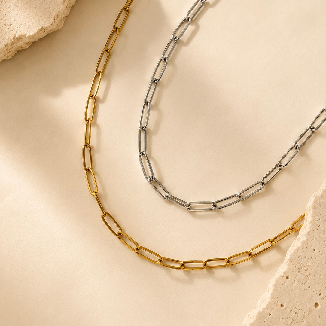 Two paperclip chain necklaces in gold and silver tones, arranged on a warm beige textured surface with stone accents.