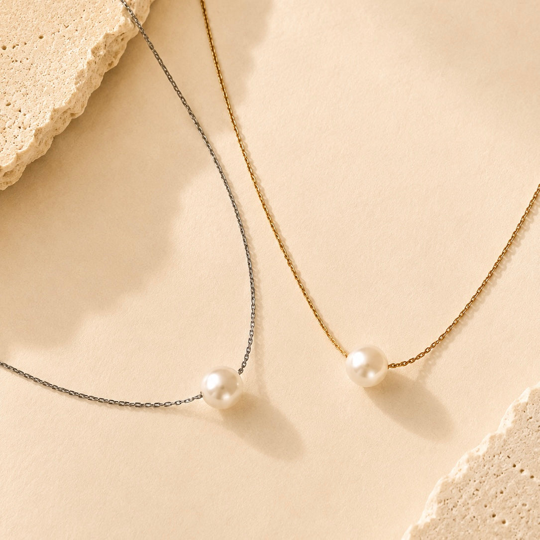 Two delicate necklace chains laid on a warm beige surface: one silver-toned chain with a white pearl pendant and one gold-toned chain with a white pearl pendant. Decorative textured stone pieces frame the corners.