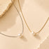 Two delicate necklace chains laid on a warm beige surface: one silver-toned chain with a white pearl pendant and one gold-toned chain with a white pearl pendant. Decorative textured stone pieces frame the corners.