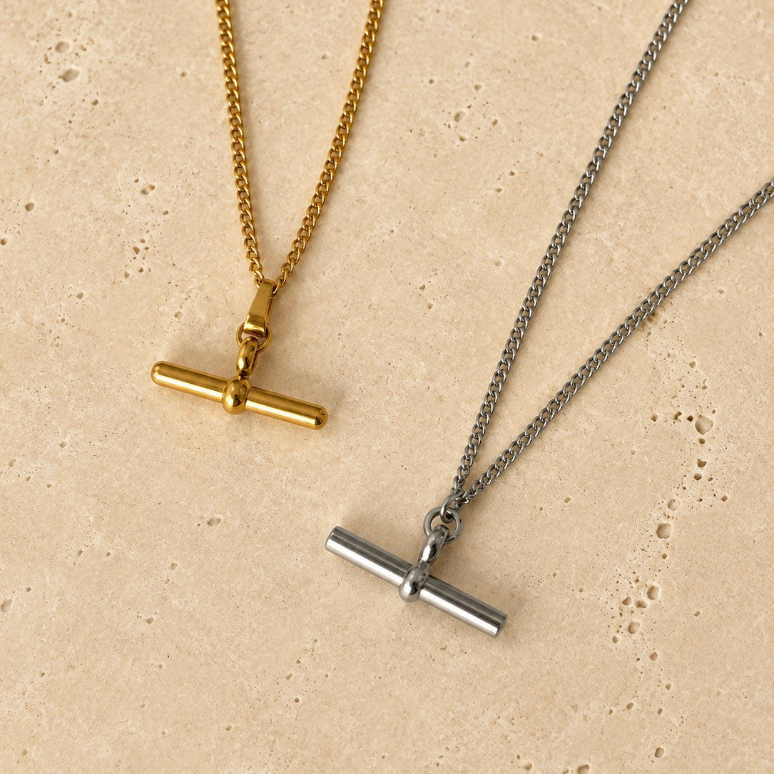 Two minimalist toggle bar necklaces displayed on a textured beige stone surface. One necklace features a polished gold-toned toggle bar on a gold chain, and the other features a silver-toned toggle bar on a silver chain.