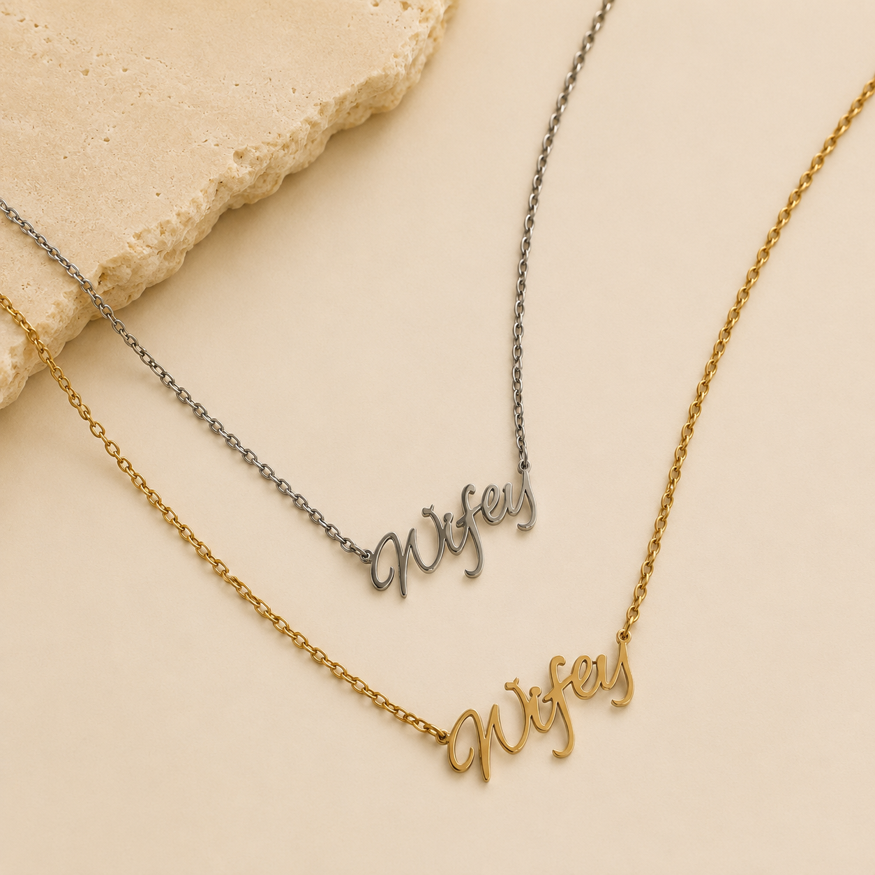 Two personalized name necklaces with cursive script pendants reading 'Nysef', one in gold and one in silver, displayed on a beige background with a stone prop.